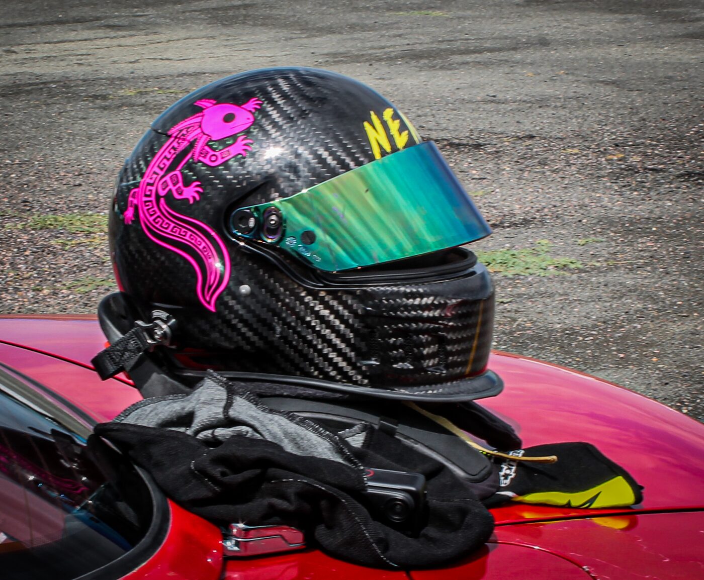 A carbon-fibre racing helmet with the pink axolotl logo painted on the side, resting on the roof of a red car